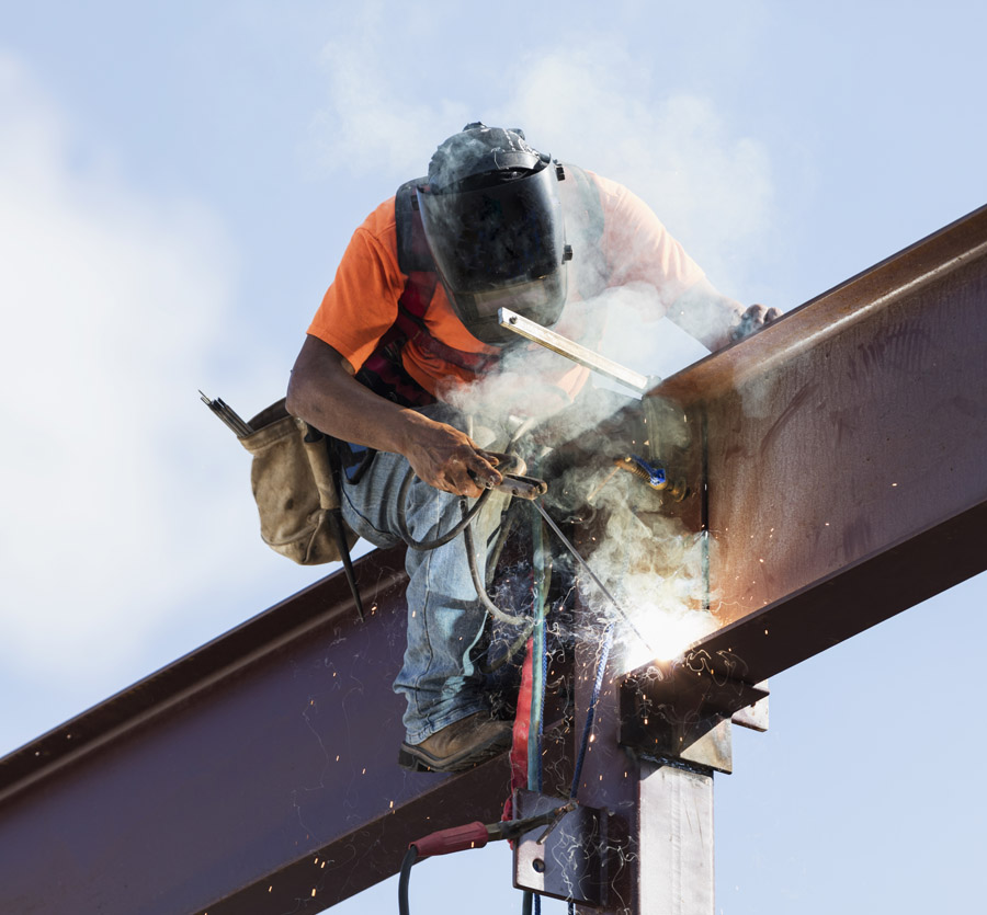 A man sits atop a large beam welding the joints.