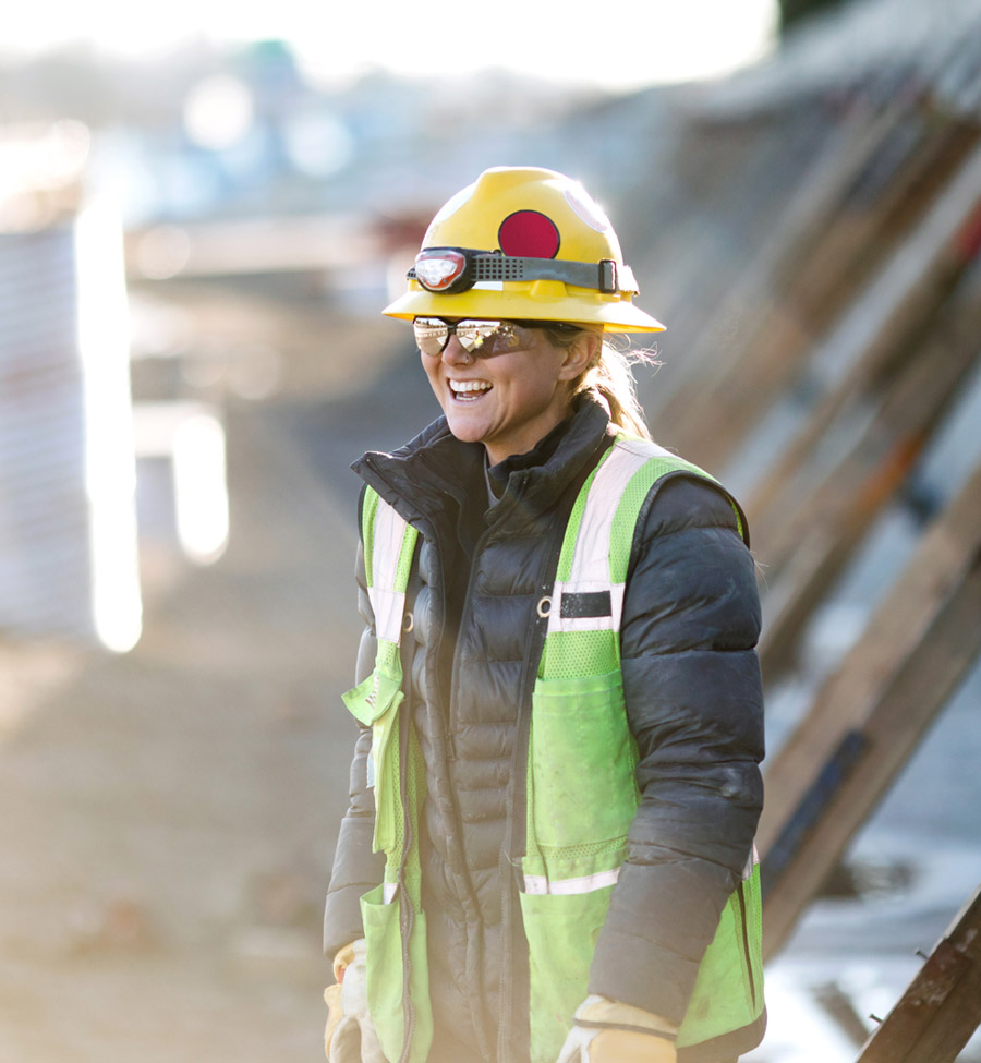 A woman wearing a neon vest and construction hat smiling on a construction site.
