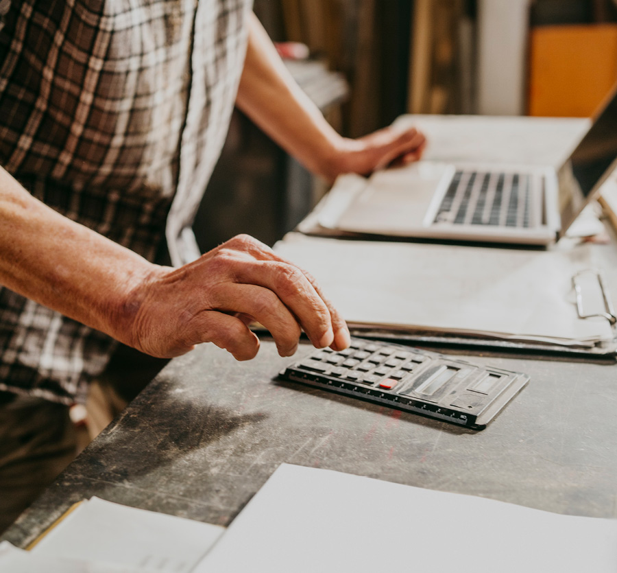 Man types on a calculator with open laptop and clipboard of papers nearby.