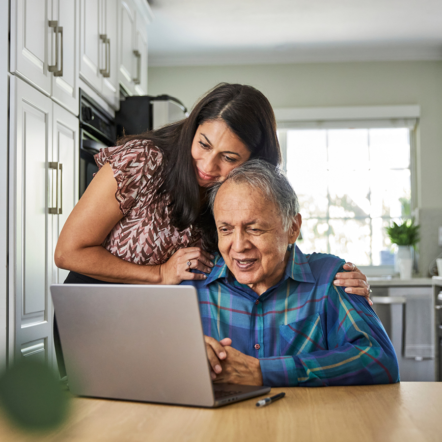Grandfather and daughter gather around the kitchen table to look at a laptop screen.