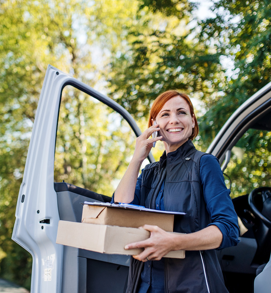 A smiling women stands beside open car, holding packages in one hand with a phone to her ear.