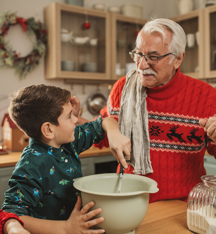 Grandfather in festive red Christmas sweater in the kitchen with young grandson doing some holiday baking together.