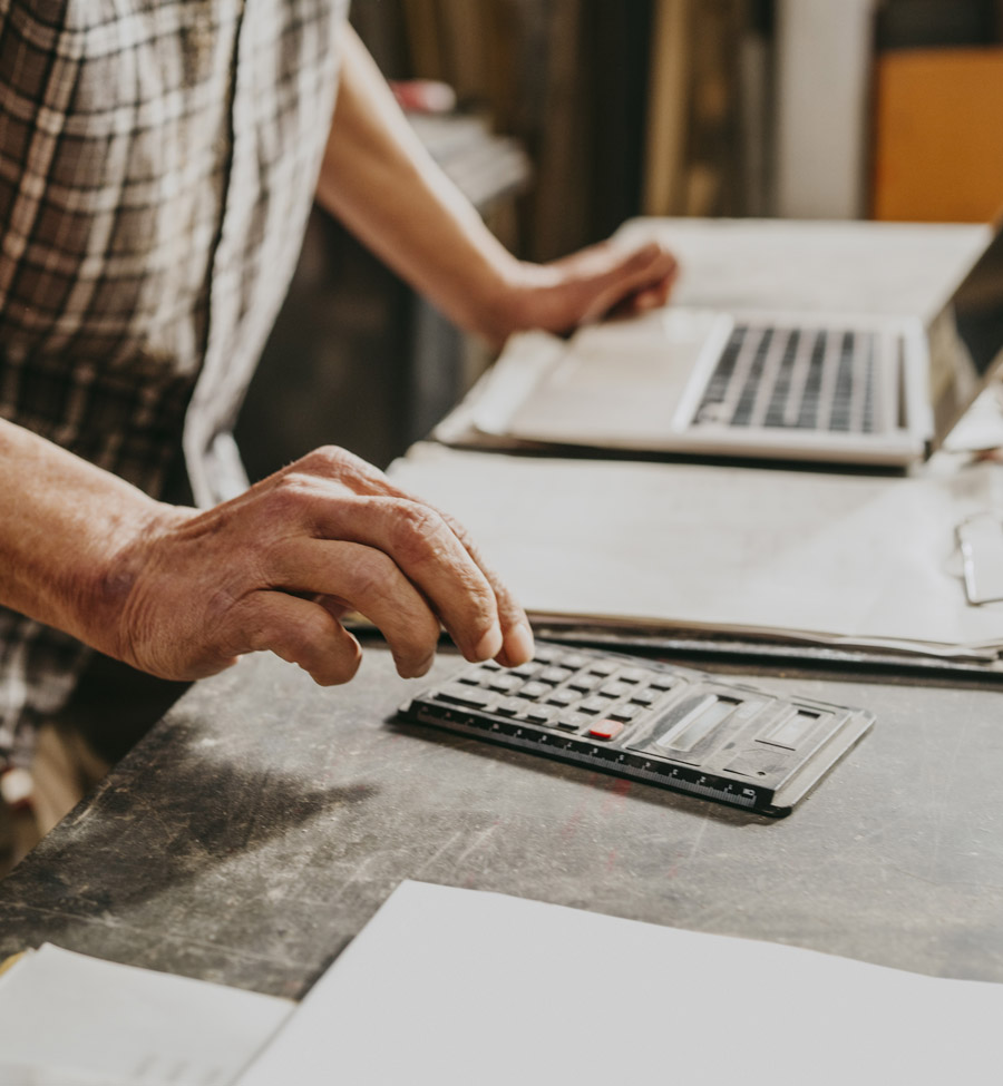 Man types on a calculator with open laptop and clipboard of papers nearby.
