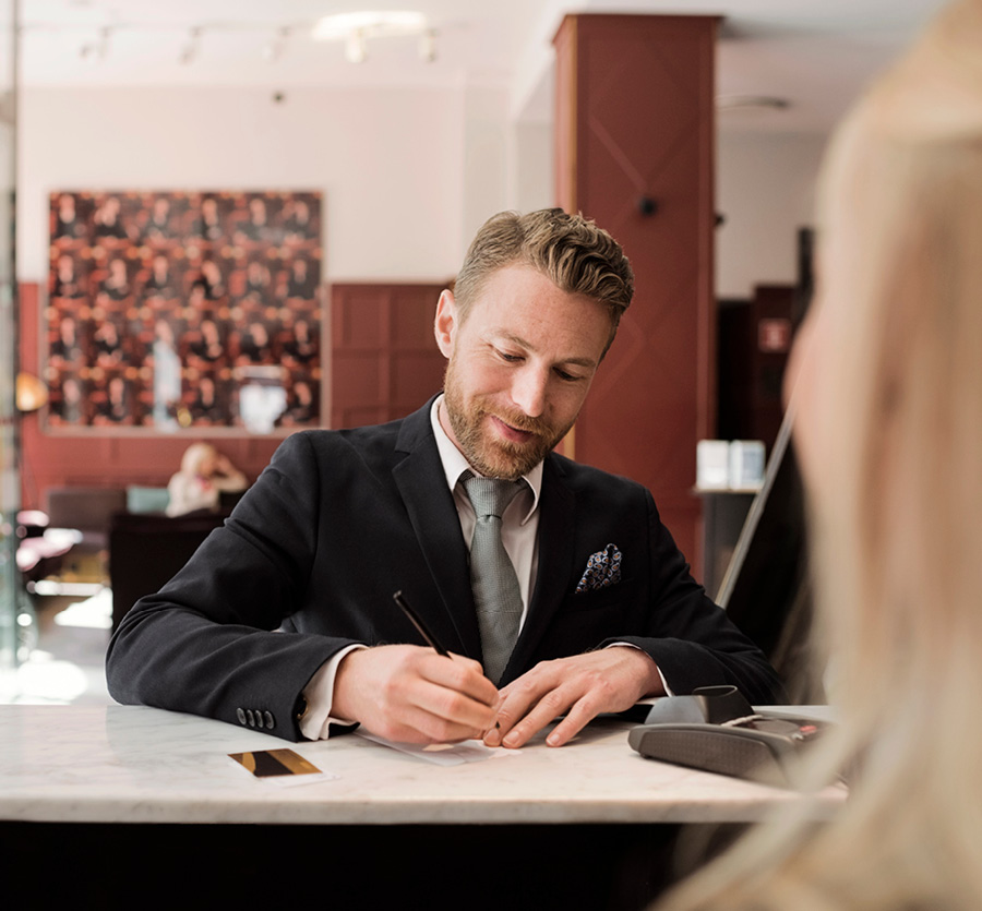 A man in a tie stands at a tall counter writing a note for a customer.