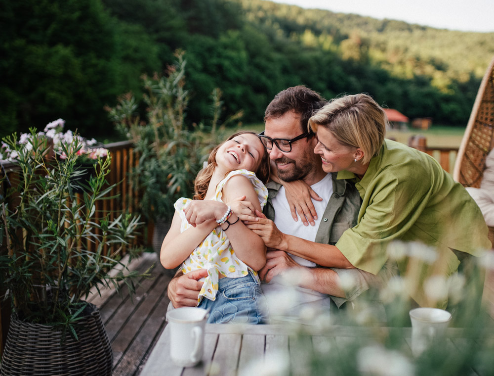 A mom and dad hugging their daughter while seated on a deck overlooking a yard.