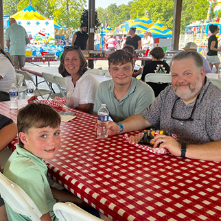 Family enjoys a meal at a picnic table a community event. They all smile for the camera. 