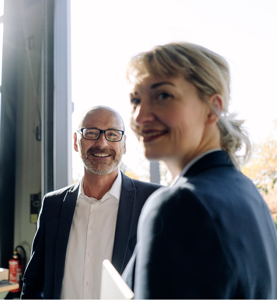 Two business colleagues stand together smiling, looking towards the camera.