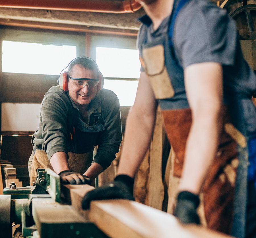 Two men in a workshop plane a wooden board together.