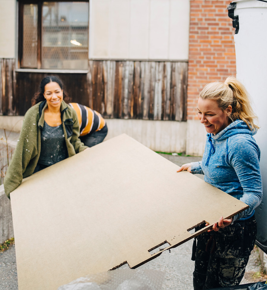 Two women maneuver a large board behind a warehouse.