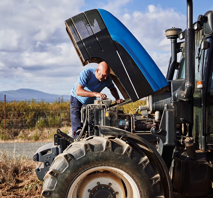 A man stands working under the hood of a tractor in a field with rolling mountains in the distance.