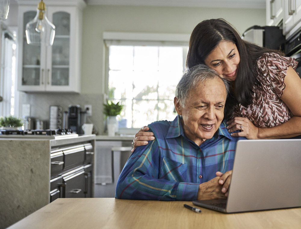 A man and woman hug while looking at a laptop screen at the kitchen table.